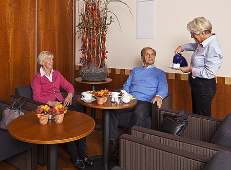 Das Bild zeigt drei ältere Menschen. Eine Frau davon, schüttet den beiden anderen eine Tasse Kaffee ein